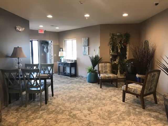 Interior view of a common area in a senior living facility with carpeted floor, several chairs arranged in small groups, a table with chairs on the left, potted plants, wall decorations, and a door leading outside with an exit sign above it.