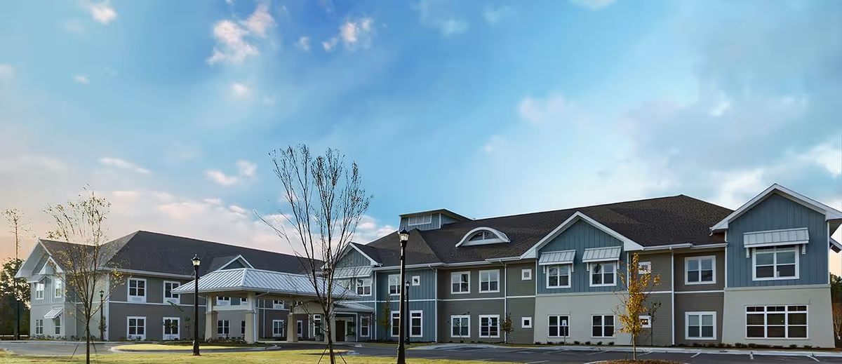 Front exterior of a two-story senior living building with a covered porte-cochère, landscaped grounds, and a blue sky.