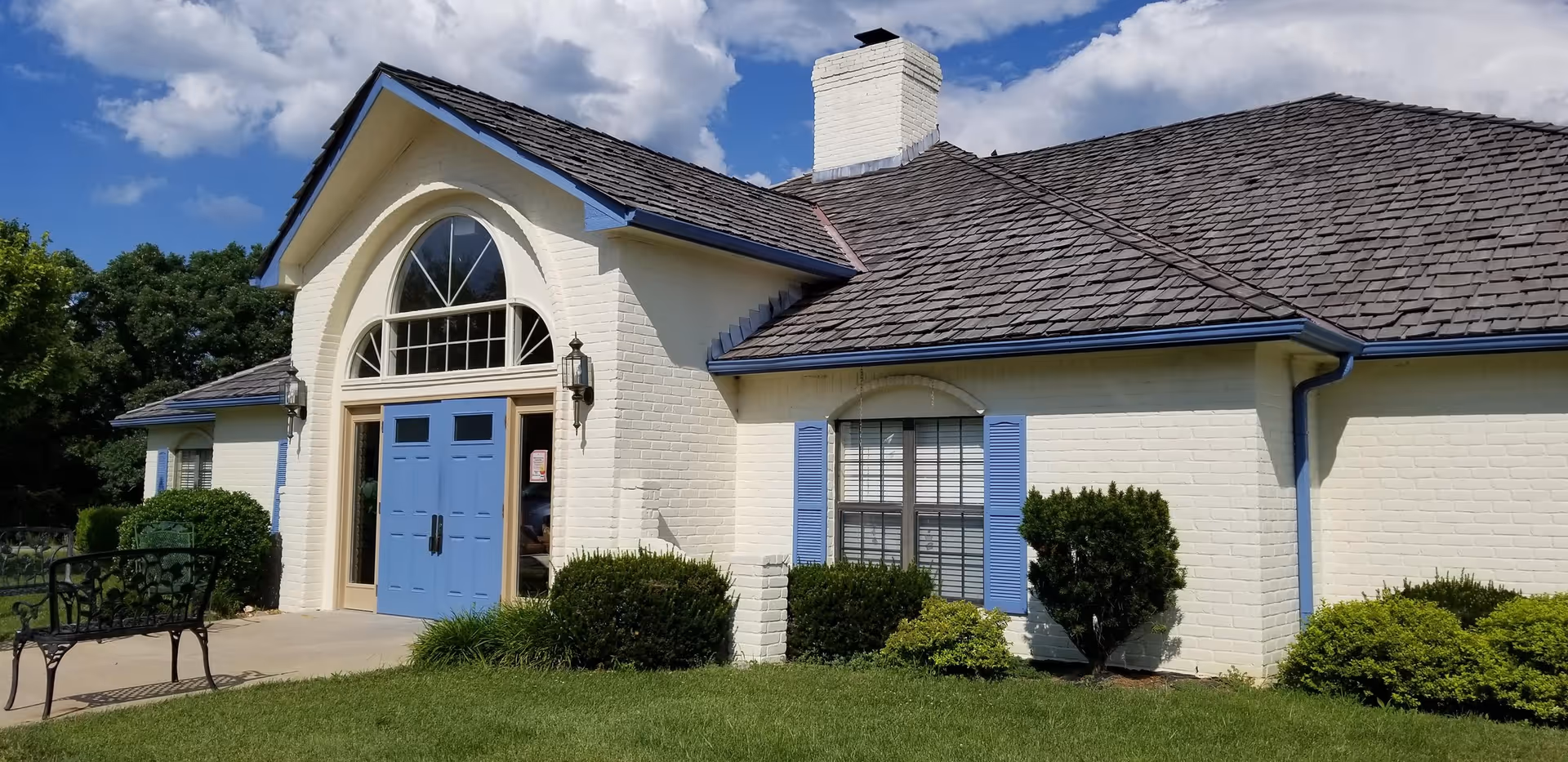 Front exterior of a light-colored brick building with blue double doors, an arched window above, blue shutters, and landscaped shrubs.