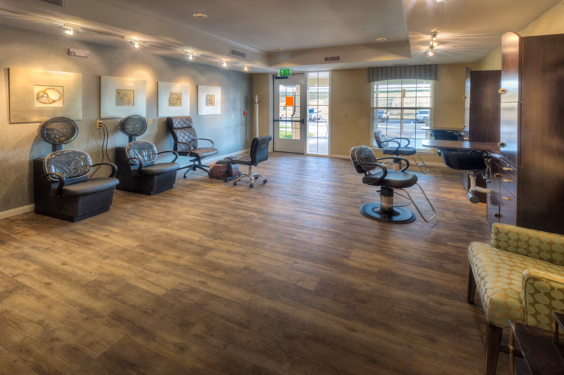 Interior view of a salon area in an assisted living facility with several black salon chairs, hair dryers, and a large window letting in natural light. The room has wooden flooring, light-colored walls with framed artwork, and a comfortable patterned armchair in the corner.