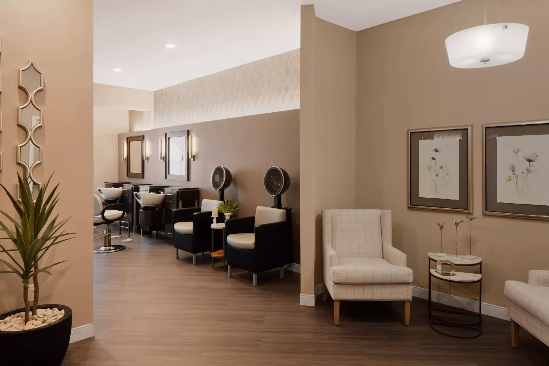 Interior view of a salon area in a senior living facility with beige walls and wood flooring. The space includes salon chairs with hair dryers, washbasins, mirrors, and a seating area with two beige armchairs and a small round table. Decorative wall art and a potted plant are also visible.