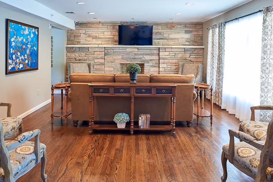 A cozy living room with a brown sofa facing a stone fireplace with a mounted flat-screen TV above it. There are two patterned armchairs on either side of the fireplace, a wooden console table behind the sofa with a potted plant and books, and two small side tables next to the sofa. The room has wooden flooring, light-colored walls, a large window with patterned curtains, and a framed painting of a blossoming tree on the left wall.