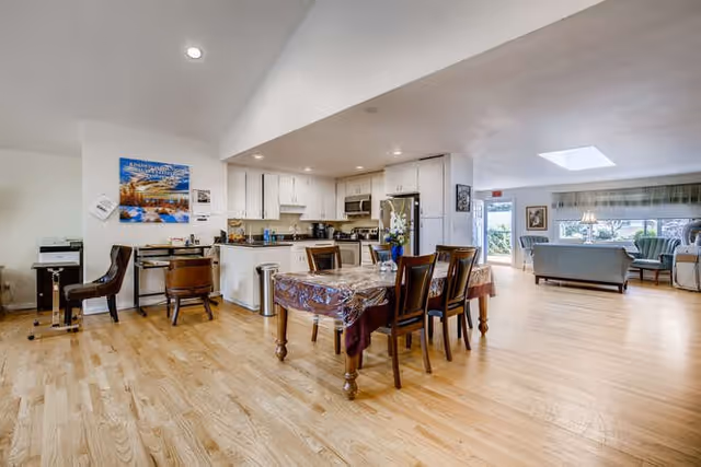 Spacious open-concept interior featuring a dining area with a wooden table covered by a plastic tablecloth and six chairs, adjacent to a modern kitchen with white cabinets and stainless steel appliances. The room has light hardwood flooring and a vaulted ceiling. In the background, there is a living area with sofas and large windows letting in natural light.