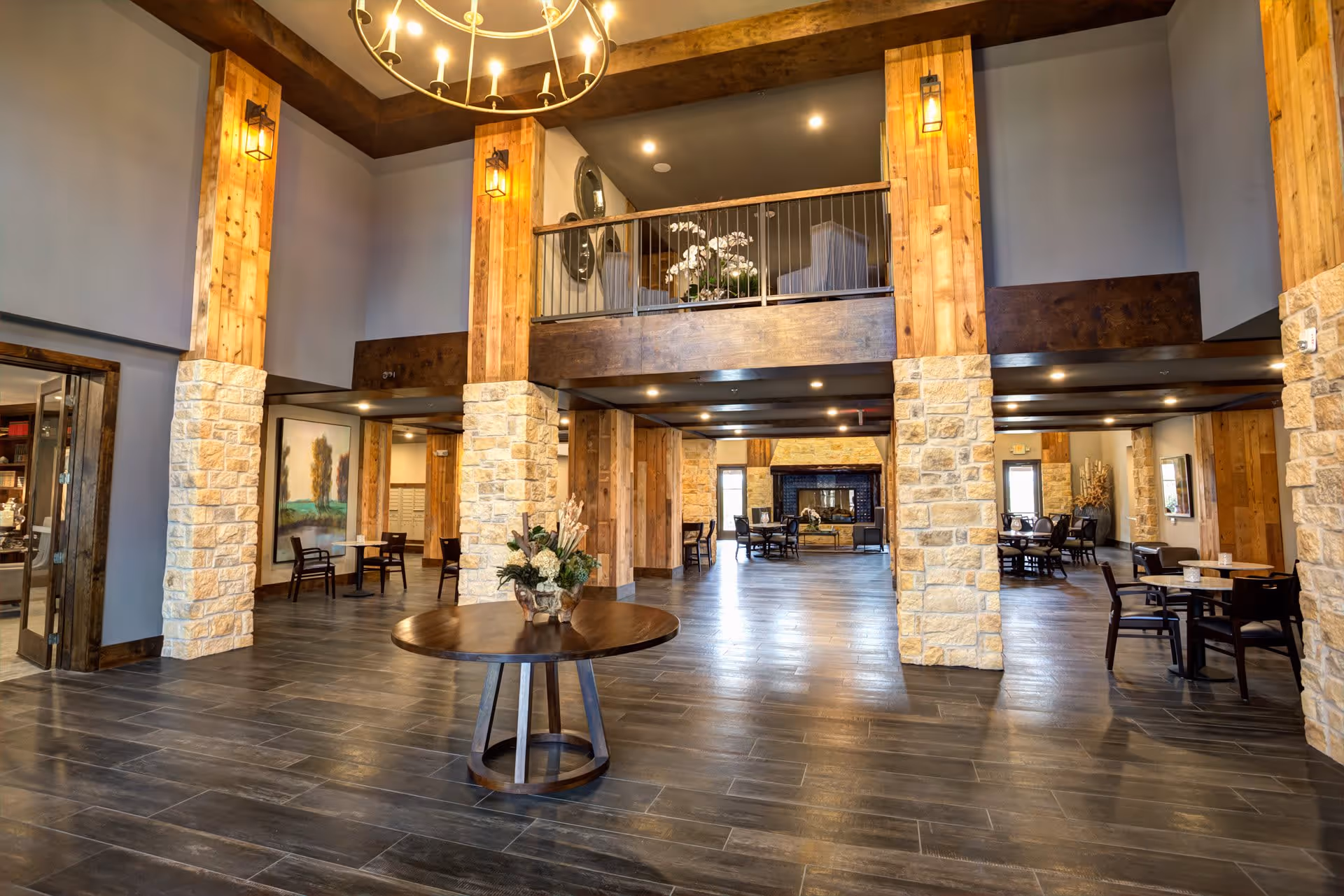 Spacious senior living facility common area with high ceilings, wooden beams, stone pillars, and a round wooden table with a floral arrangement in the center. Multiple seating areas with tables and chairs are visible, along with a fireplace in the background and a balcony with additional seating above.