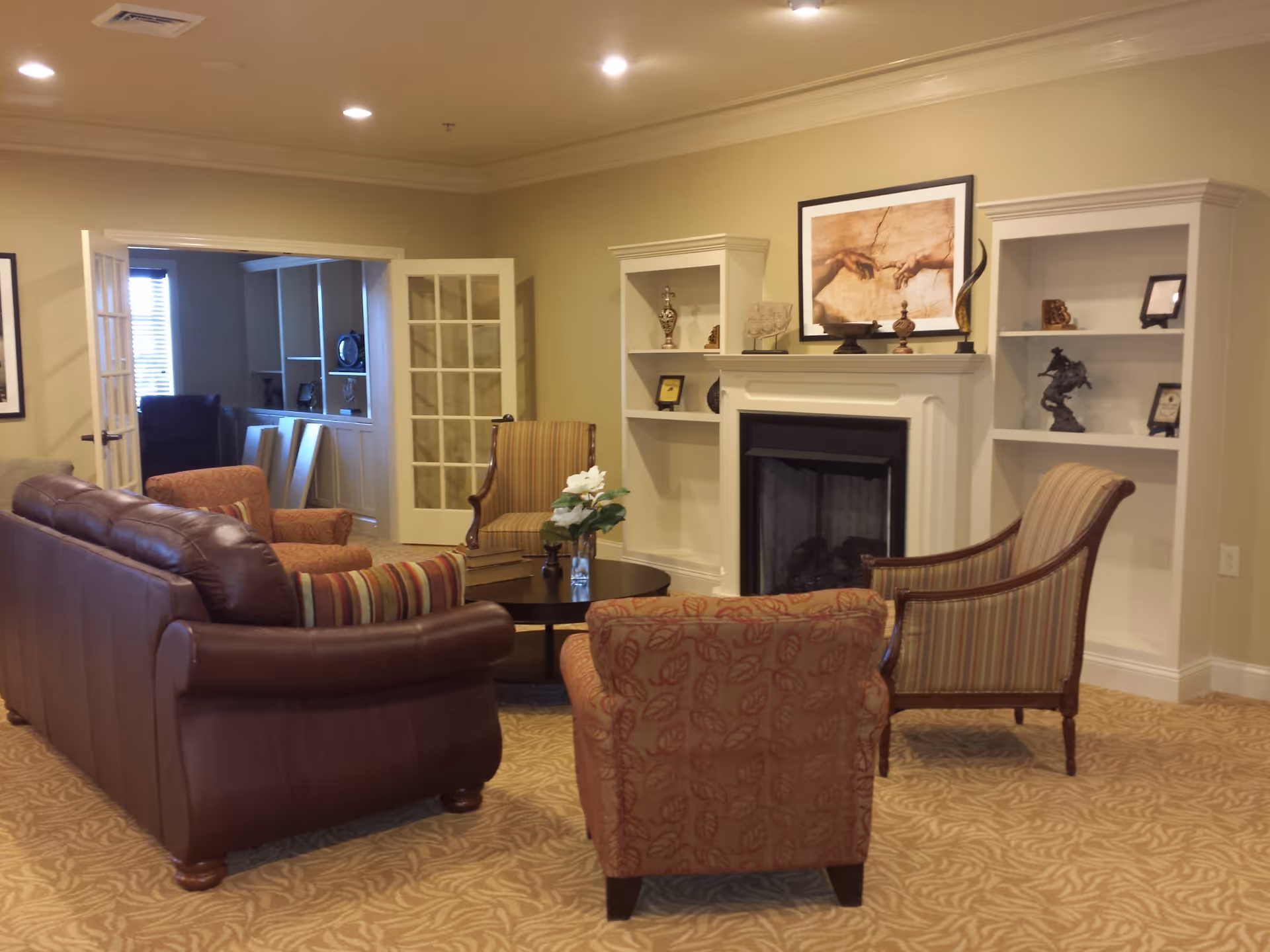 A cozy living room with a brown leather sofa, two patterned armchairs, and a striped armchair arranged around a round coffee table with a vase of white flowers. The room features a fireplace with built-in white shelves on either side, decorated with various ornaments and a framed artwork above the mantel. French doors lead to another room with additional seating and shelving.