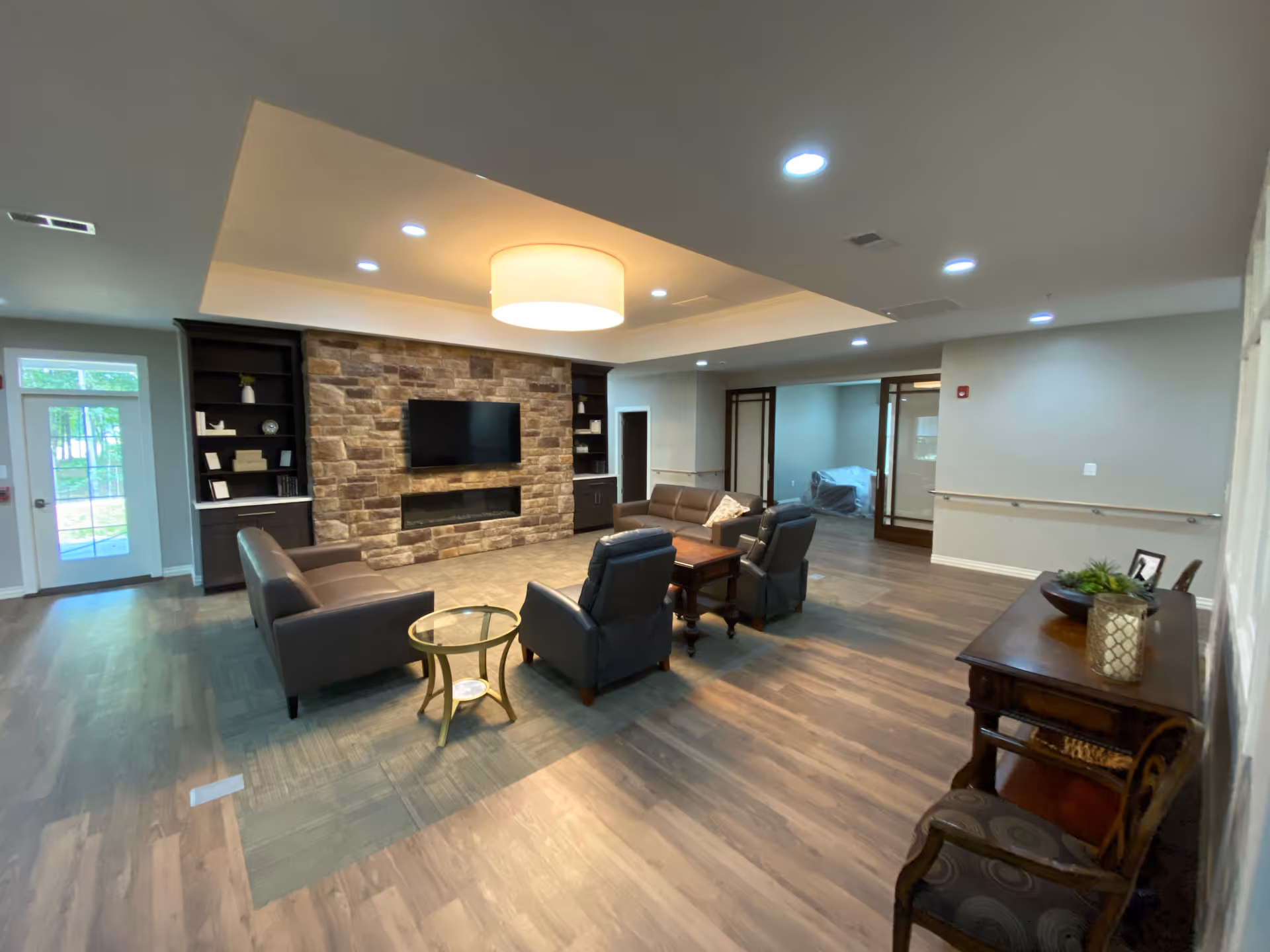 A spacious senior living common area with a stone accent wall featuring a mounted flat-screen TV and a built-in electric fireplace. The room has comfortable seating including two leather sofas and two armchairs arranged around a wooden coffee table. There are built-in shelves on either side of the stone wall with decorative items. The flooring is a combination of wood and carpet, and the ceiling has recessed lighting and a large central light fixture. A wooden console table with decorative items and a chair are positioned against the right wall. Glass doors and windows provide natural light from the left side of the room.
