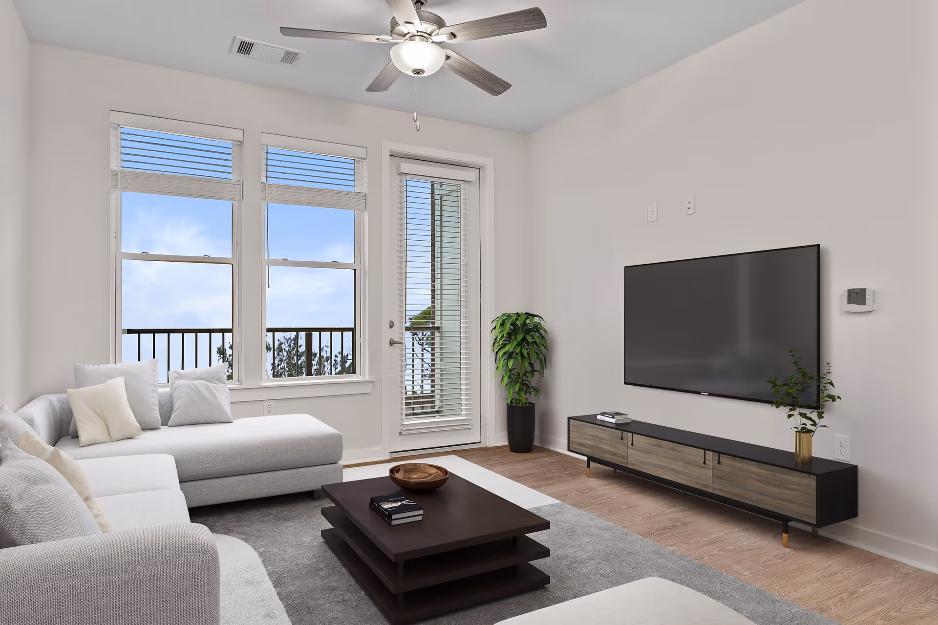 Bright neutral-toned living room with a white sectional sofa, dark wood coffee table, wall-mounted TV, and windows/door opening to a balcony.