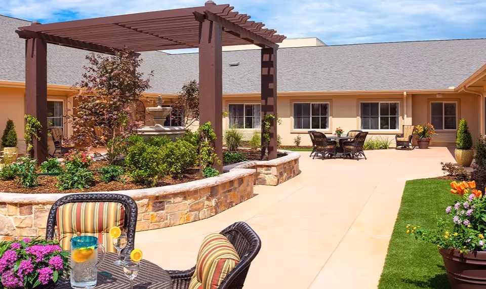 Outdoor courtyard area at Meadowbrook Memory Care Community featuring a stone planter with greenery and a fountain under a wooden pergola, surrounded by patio seating with tables and chairs, colorful flowers, and a well-maintained lawn under a partly cloudy sky.