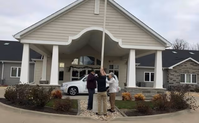 Three elderly people standing together outside the entrance of a building with a covered driveway, holding onto a flagpole. The building has beige siding, white columns, and a stone base with a white vehicle parked nearby.