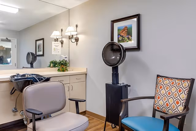 Interior view of a hair salon area in a senior living facility featuring a salon chair in front of a hair washing station with a large mirror, a black hair dryer hood on a stand, a patterned cushion on a wooden chair, wall-mounted lamps, framed pictures, and a countertop with cabinets and a small plant.