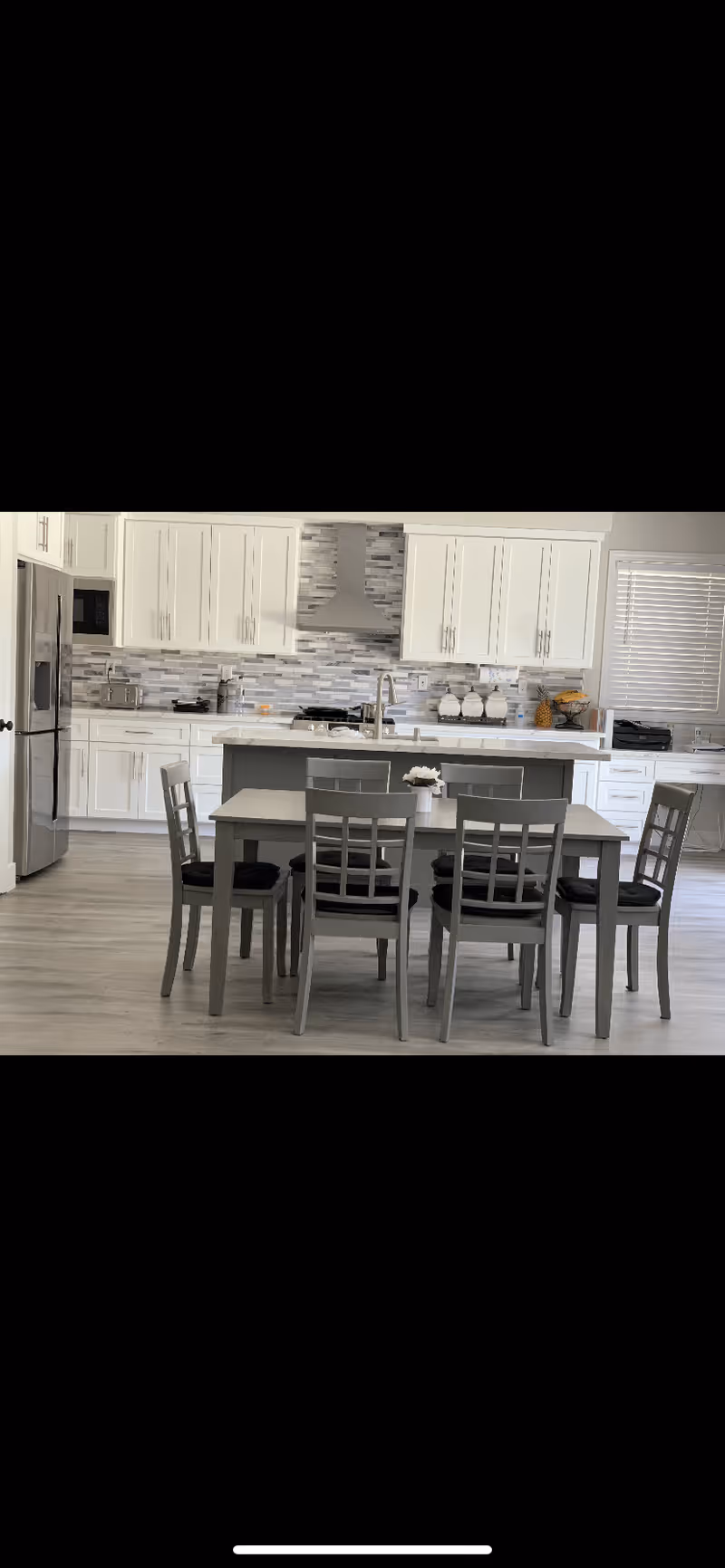 Modern kitchen and dining area with white cabinets, stainless steel refrigerator, and a gray dining table with six matching chairs. The kitchen features a tiled backsplash, a range hood, and a countertop with a sink and decorative items.