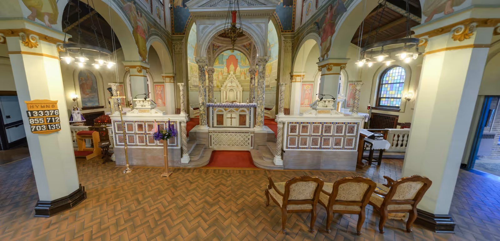 Interior view of a chapel with ornate altar, decorative columns, religious paintings on the walls and ceiling, stained glass windows, and a hymn board on the left pillar. Three upholstered wooden chairs are placed in the foreground on a tiled floor.