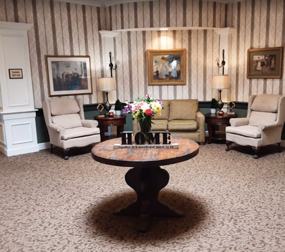 Cozy lobby-style sitting area with a round center table displaying flowers and a 'HOME' sign, flanked by armchairs and a sofa against wallpapered walls.