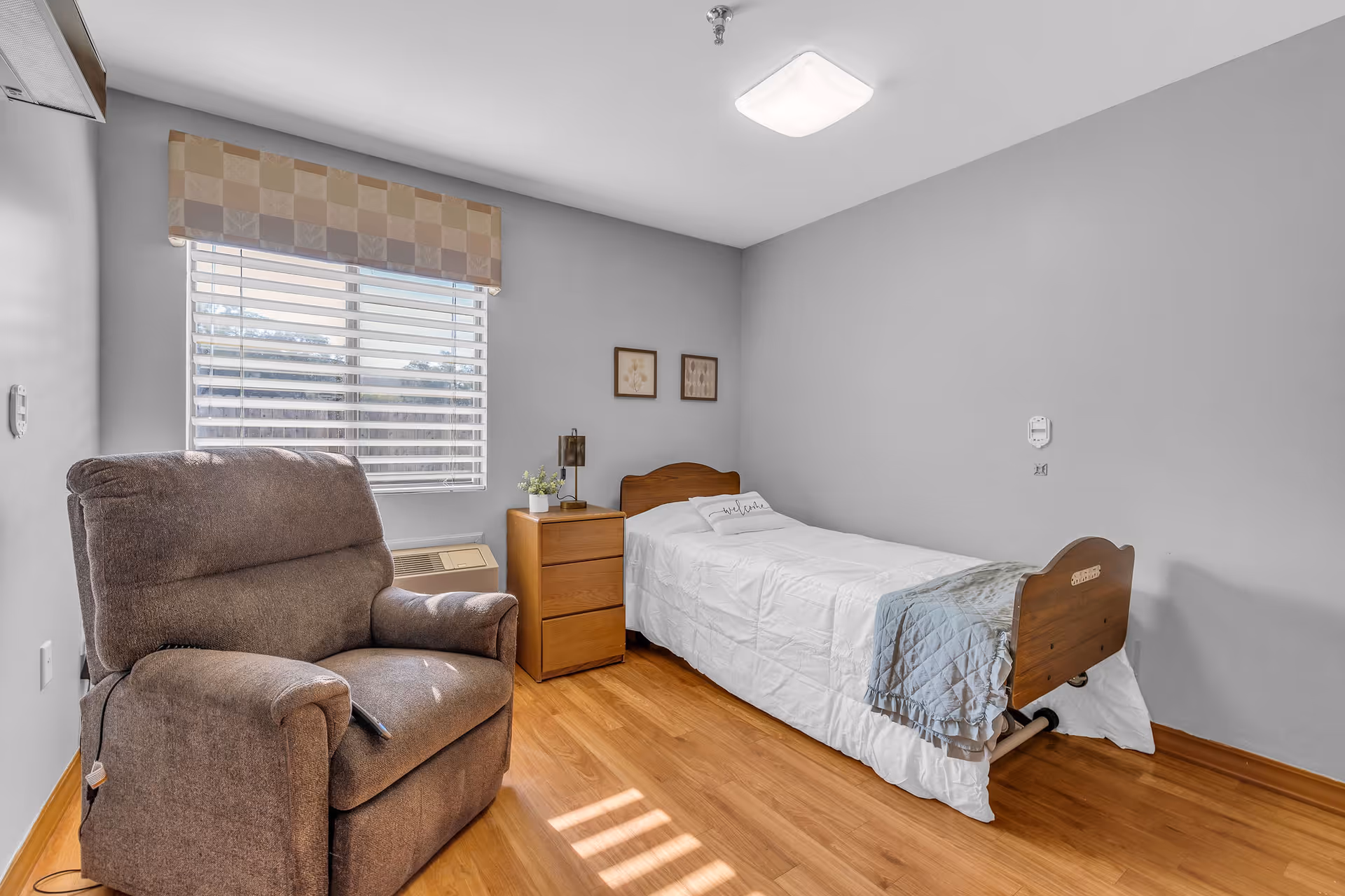 A simple bedroom with a single bed featuring a wooden headboard and footboard, white bedding, and a gray throw blanket. Next to the bed is a wooden nightstand with a small plant and a lamp. A large, cushioned recliner chair is positioned near a window with horizontal blinds and a patterned valance. The room has light gray walls and wood flooring.