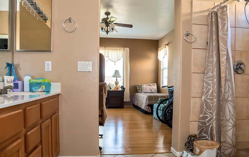View from a bathroom into a bedroom in a senior living facility. The bathroom has a sink with wooden cabinets, a mirror, and a shower with a patterned curtain. The bedroom has a bed with pillows, a nightstand with a lamp, and a wheelchair near the bed. The room has wooden flooring and windows with curtains allowing natural light.