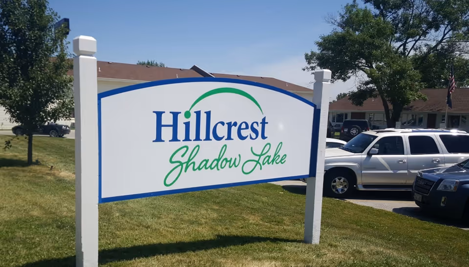 A large white sign with blue and green text that reads 'Hillcrest Shadow Lake' standing on a grassy area with trees, parked cars, and buildings in the background under a clear blue sky.