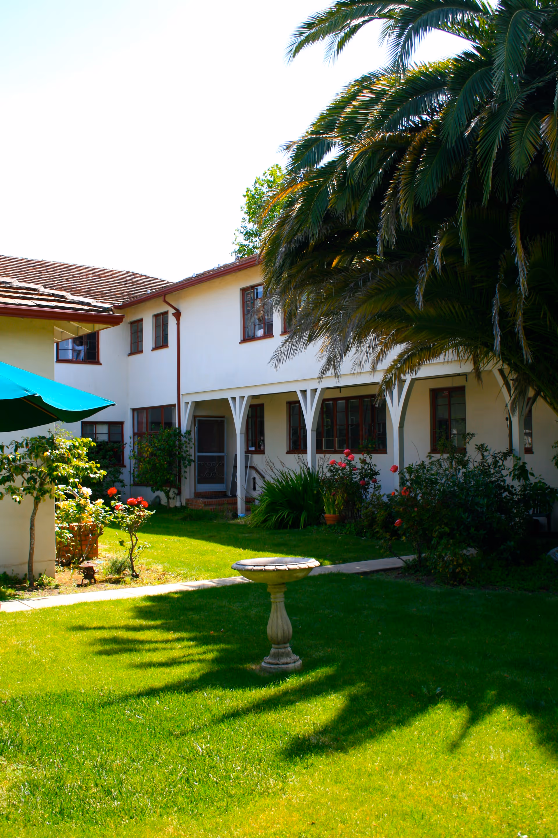 A view of Bello Gardens Assisted Living featuring a landscaped area with grass, flowers, and a birdbath in front of the building.