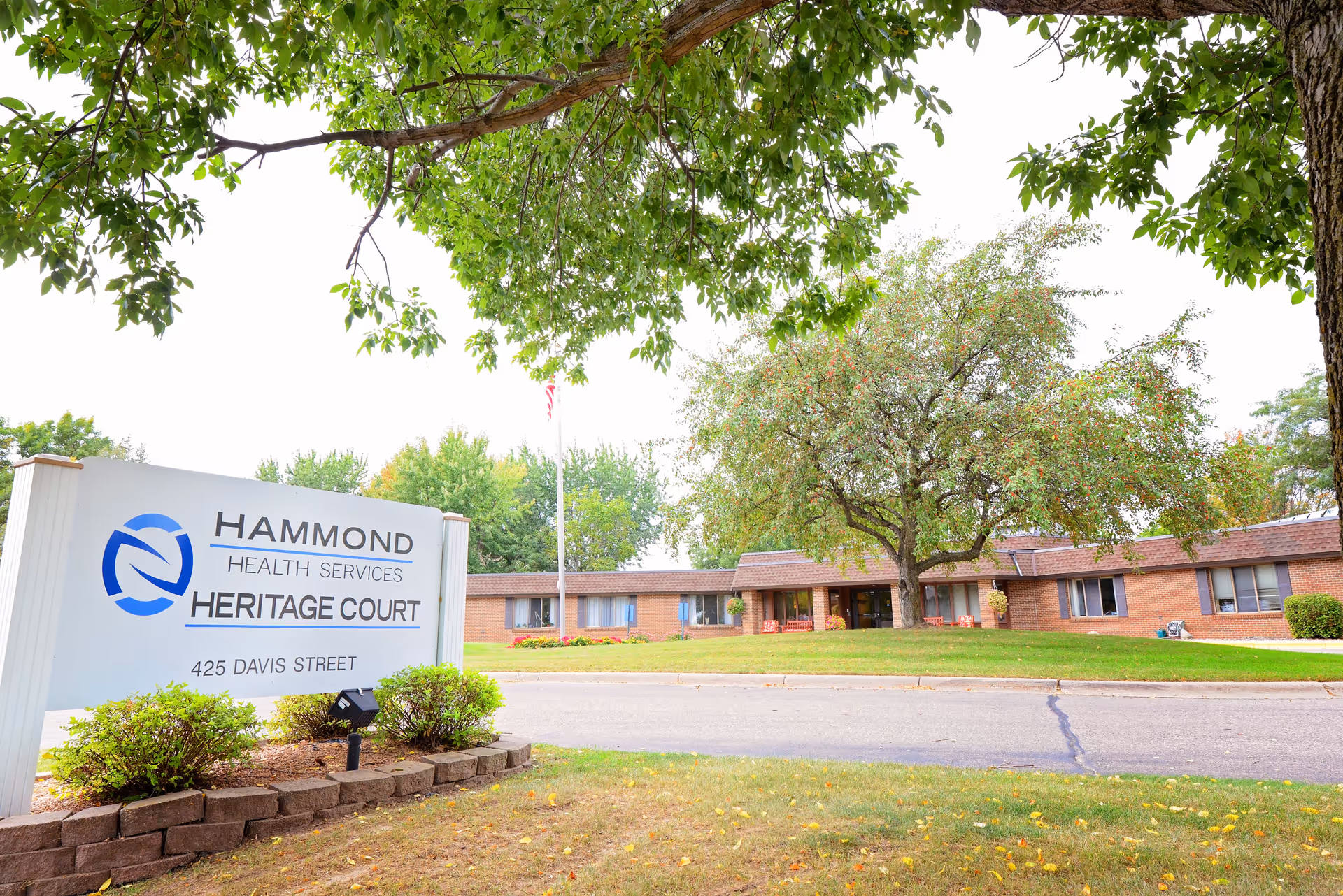 Exterior view of Heritage Court, a single-story brick building with a tree and green lawn in front. A large sign in the foreground reads 'Hammond Health Services Heritage Court 425 Davis Street'.