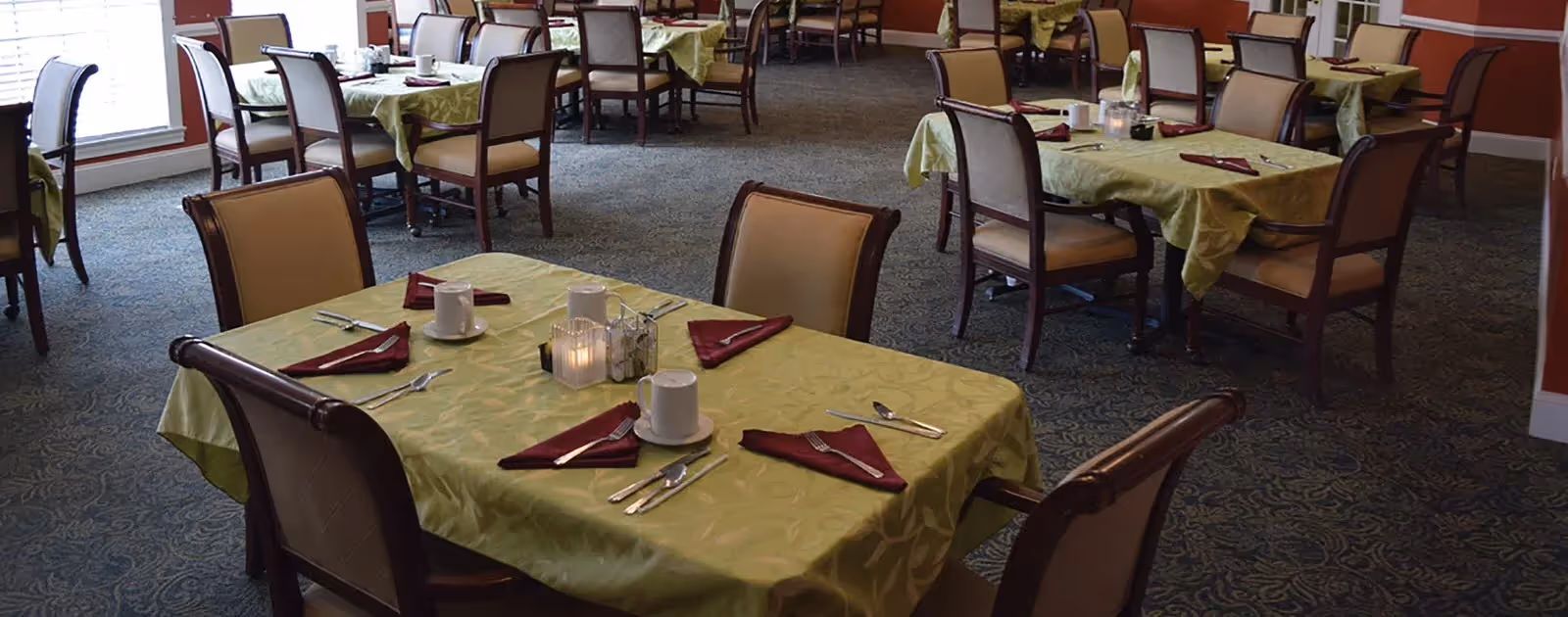 Dining room with multiple tables covered in green tablecloths, each set with white cups, silverware, and folded maroon napkins. Chairs with cushioned seats surround the tables, and large windows allow natural light into the room.
