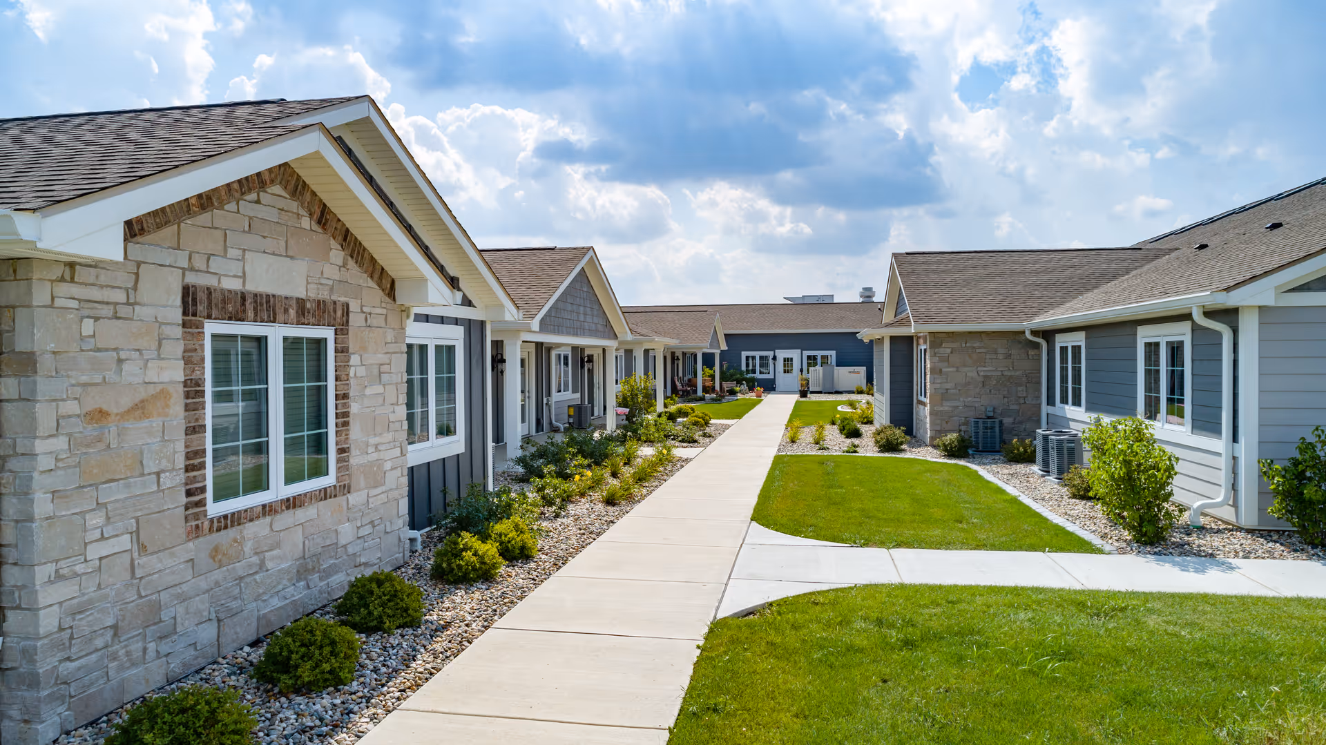 A paved walkway runs between single-story assisted living cottages with stone and siding exteriors, lawns, and landscaped shrubs under a partly cloudy sky.