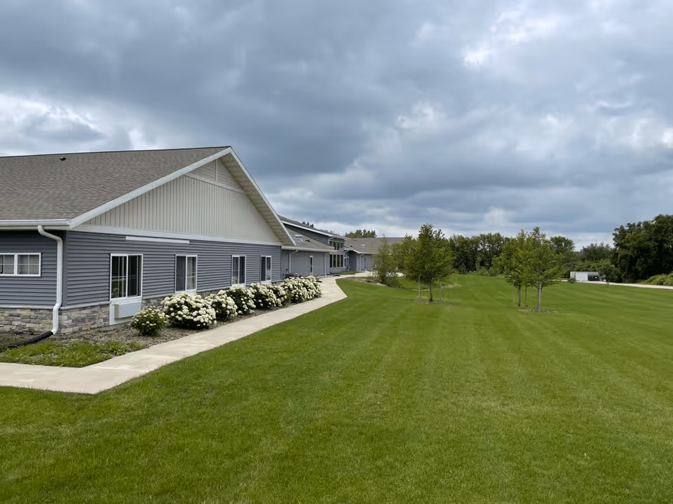 Exterior view of a single-story building with gray siding and a gable roof, surrounded by a well-maintained green lawn and small trees under a cloudy sky.