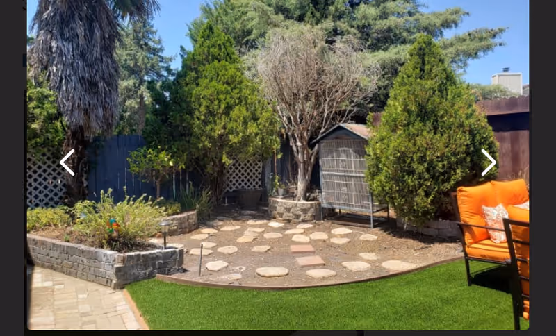Outdoor garden area with a stone pathway surrounded by raised brick flower beds, various trees and shrubs, and an orange cushioned outdoor chair on artificial grass under a clear blue sky.