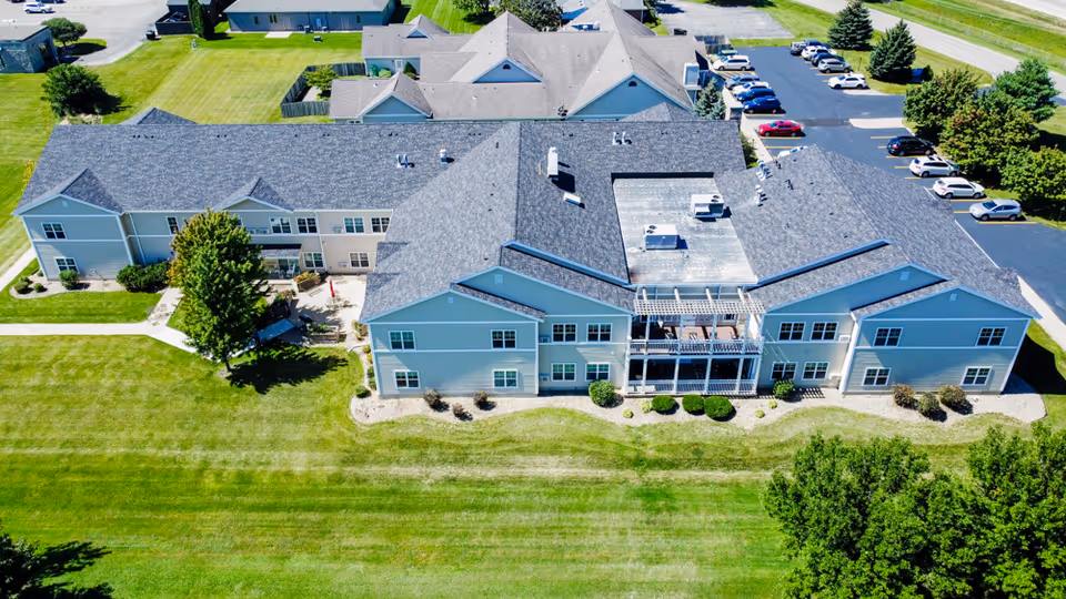 Aerial view of a senior living facility building with a gray roof and light-colored exterior walls, surrounded by well-maintained green lawns, trees, and a parking lot with several cars.