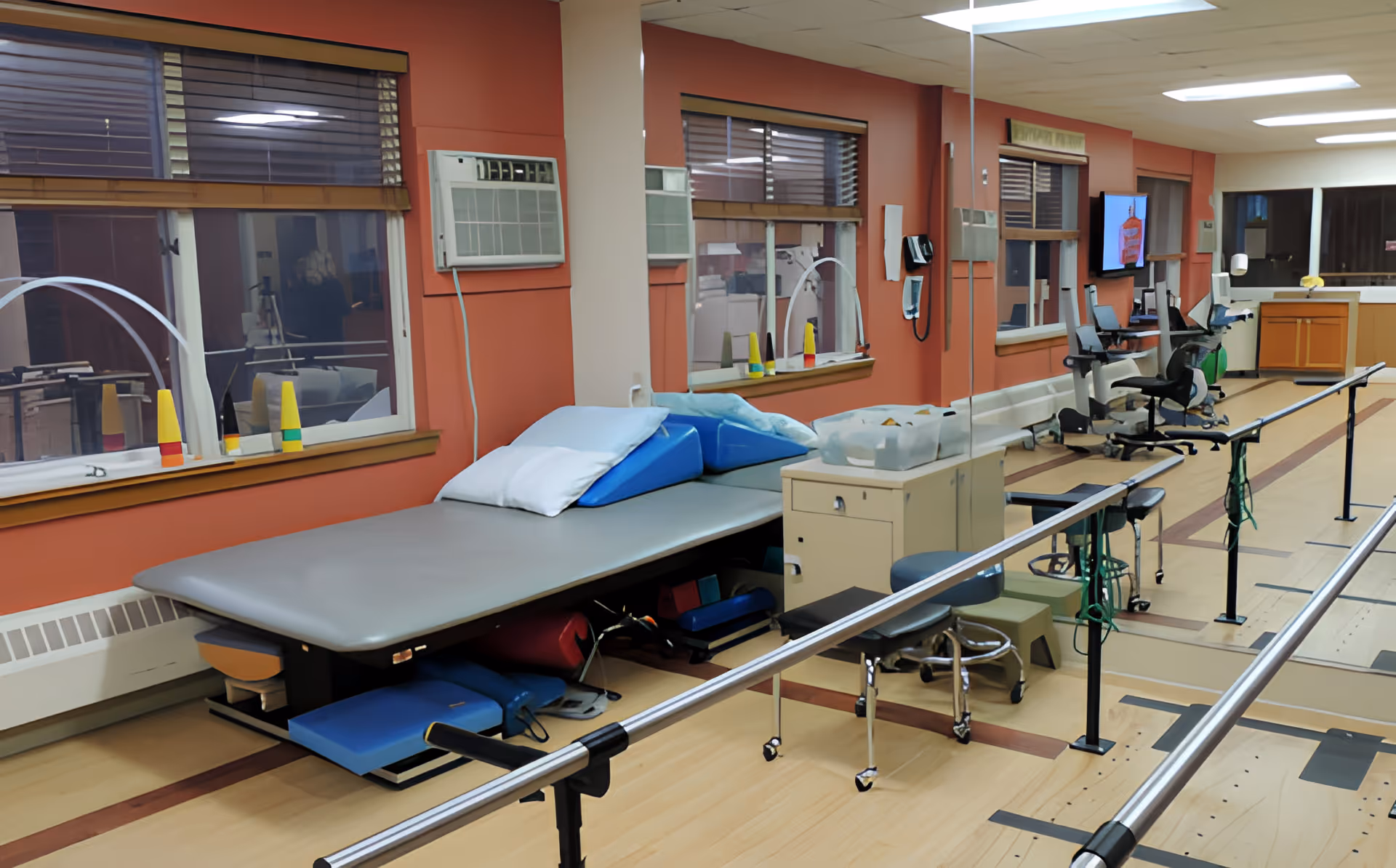 Interior view of a rehabilitation room with parallel bars for walking exercises, a padded therapy table with pillows, chairs, and various therapy equipment. The room has orange walls, windows with blinds, and a wall-mounted television.