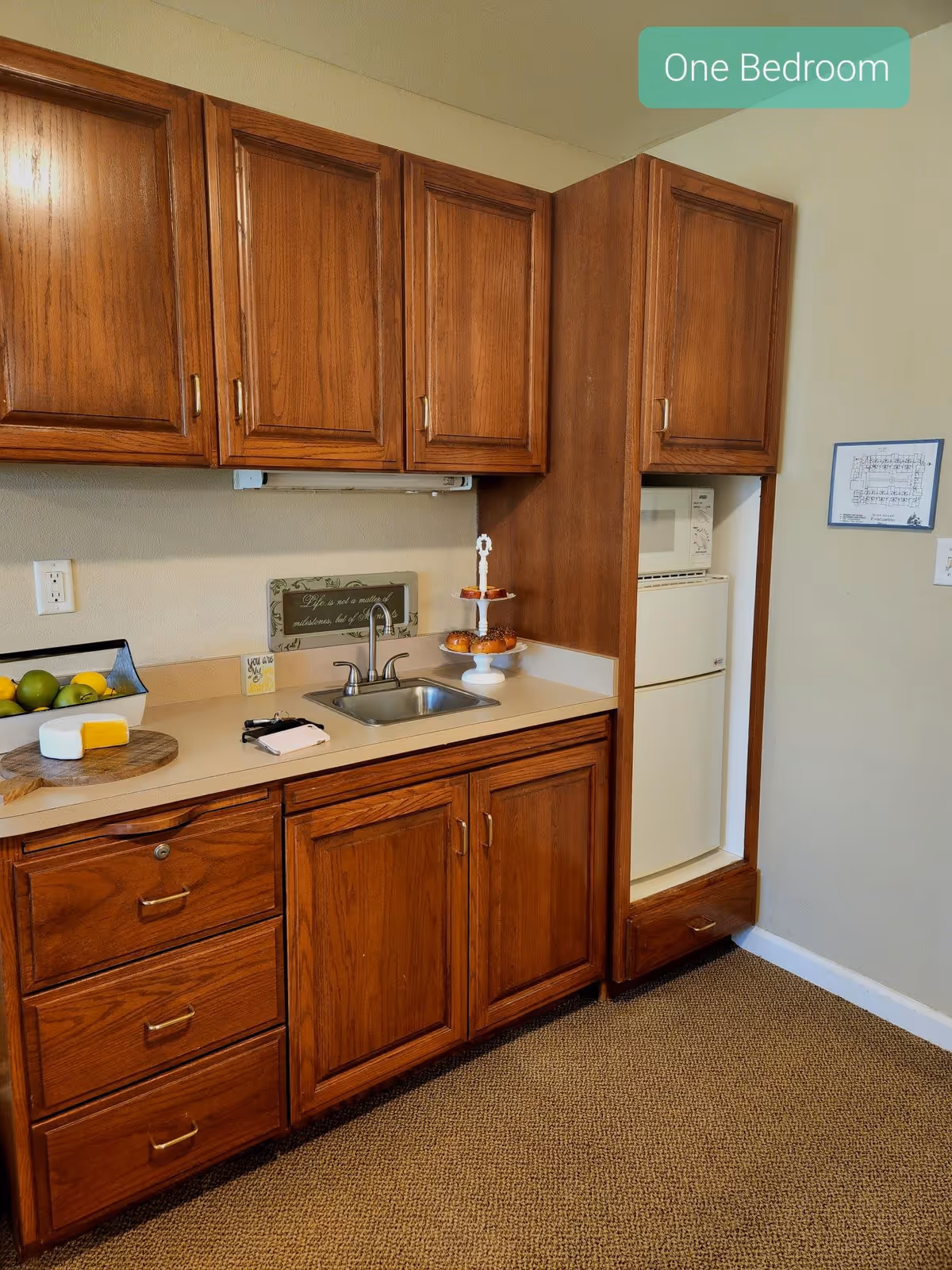 A small kitchen area with wooden cabinets, a countertop with a sink, a fruit basket, a cutting board with cheese, and a tiered tray with pastries. A microwave and mini refrigerator are built into the cabinetry. A sign on the wall reads 'One Bedroom'.