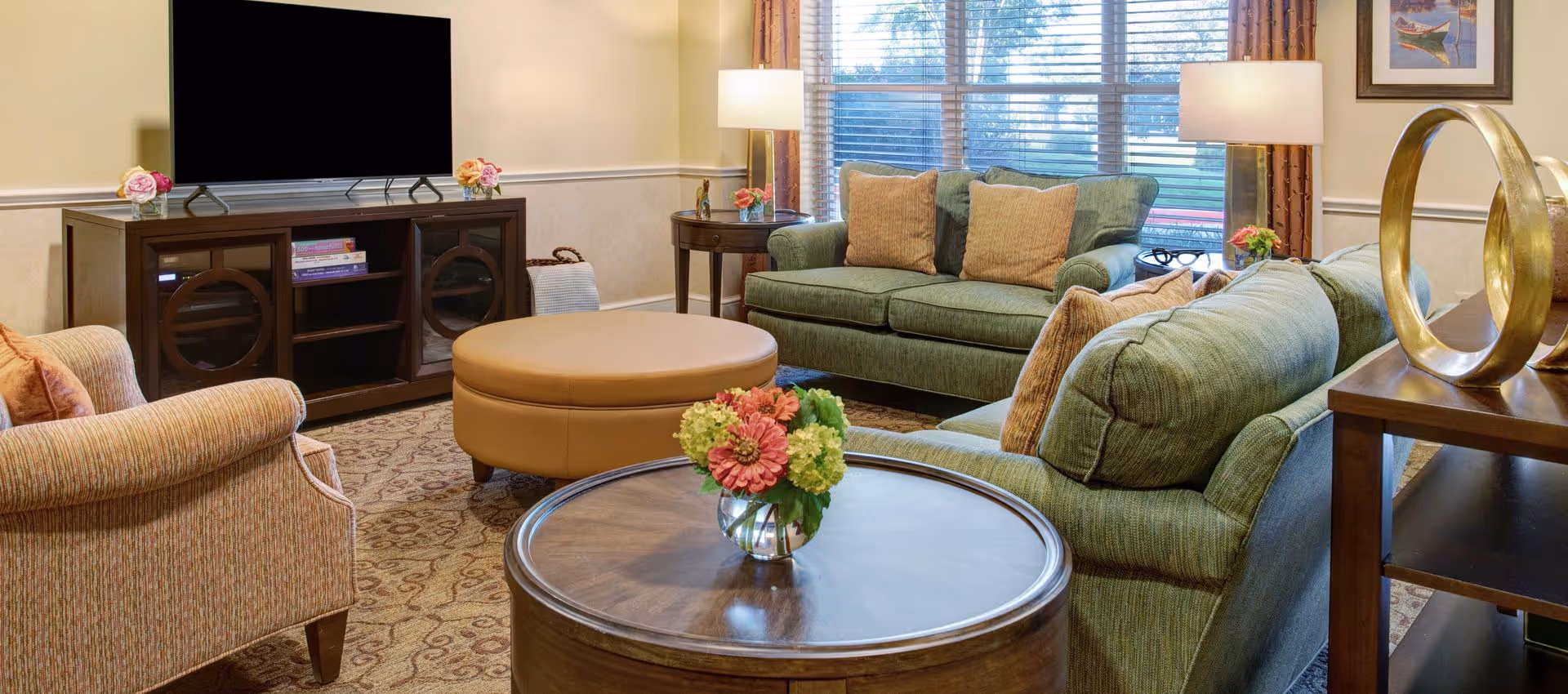 A cozy living room with two green sofas and a beige armchair arranged around a round tan ottoman and a wooden coffee table with a flower vase. A large flat-screen TV sits on a dark wooden cabinet with shelves holding books and decorative items. Two lamps with white shades are positioned near the windows with blinds, and a framed picture hangs on the wall.