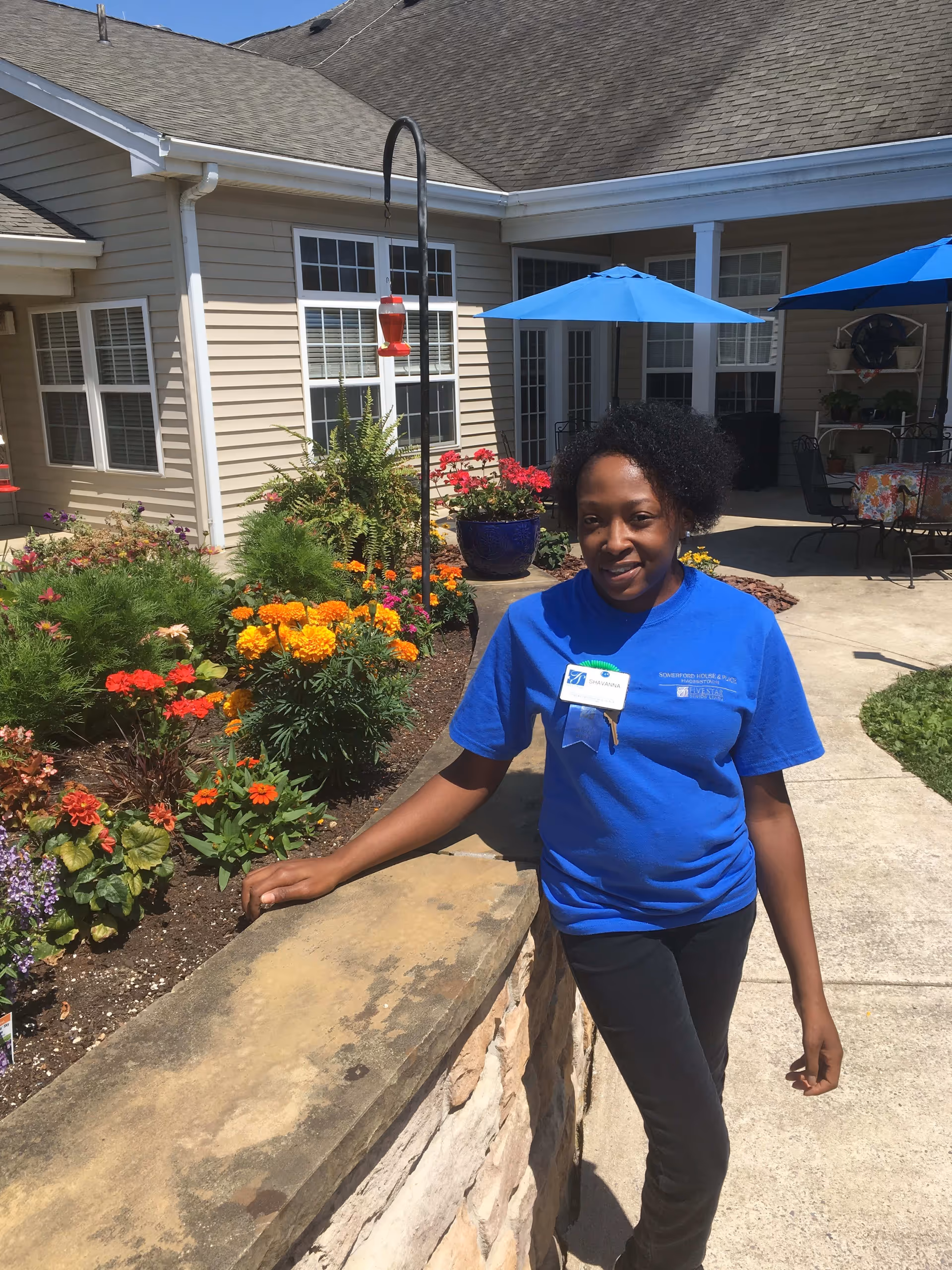 A woman wearing a blue Somerford House And Place Hagerstown t-shirt and name badge stands outdoors next to a stone wall with colorful flowers and plants behind her. The background shows a building with beige siding, windows, and blue patio umbrellas over tables.