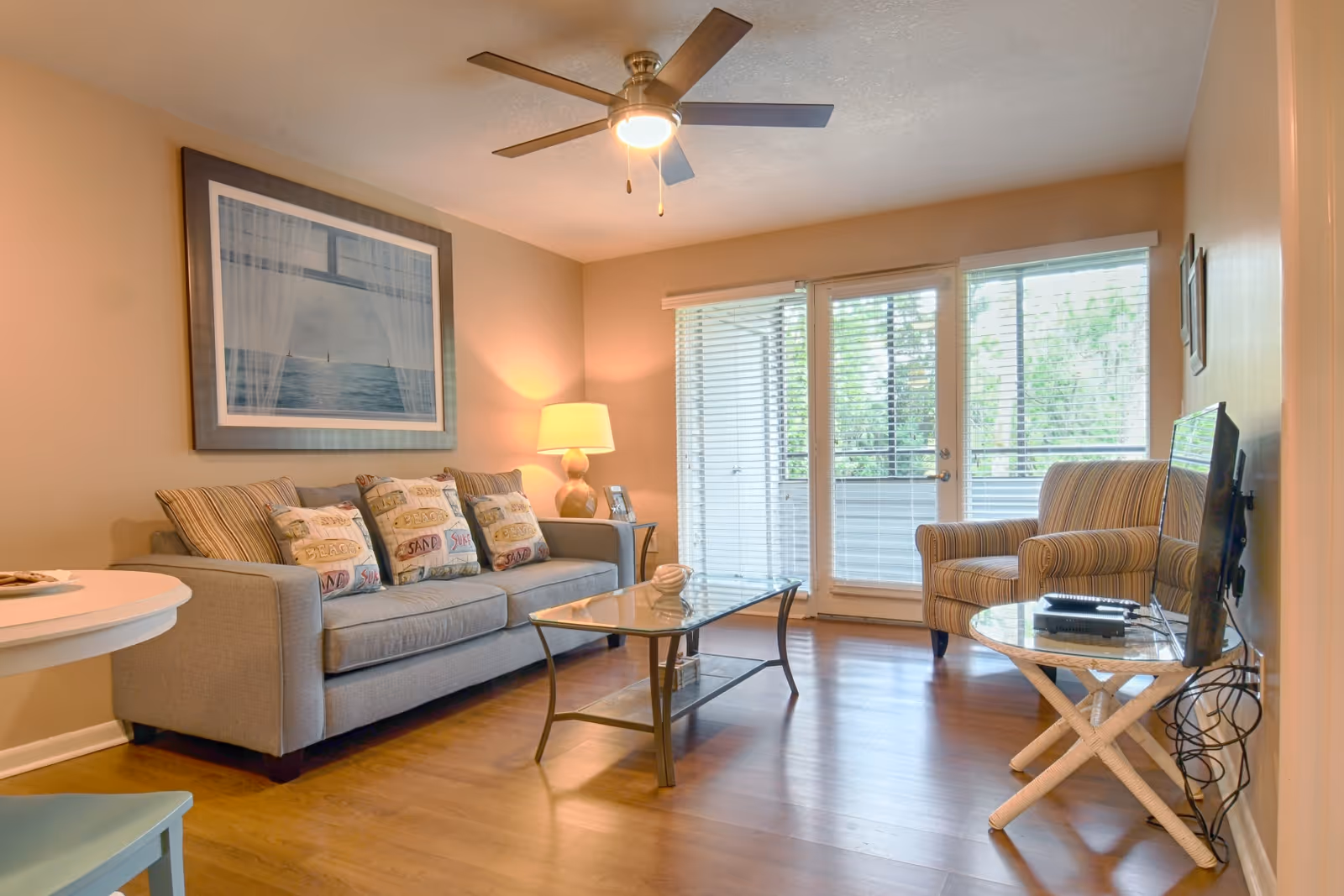 A cozy living room with a gray sofa adorned with decorative pillows, a glass coffee table, a striped armchair, a side table with a lamp, and a flat-screen TV mounted on the wall. Large windows with blinds and a glass door allow natural light to fill the room. The ceiling has a modern fan with a light.