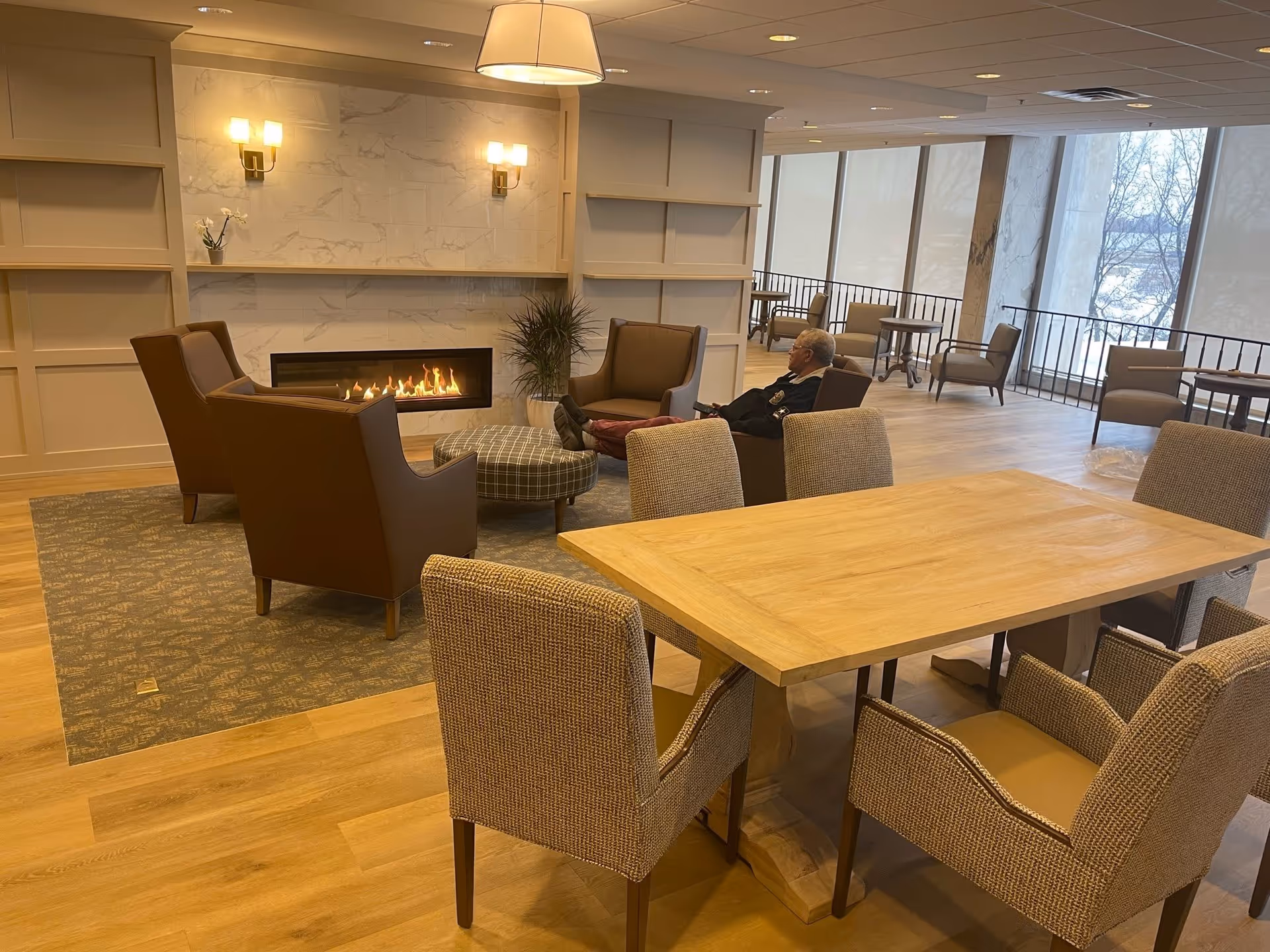 A cozy common area in a retirement community featuring a modern fireplace with a marble surround, four brown armchairs arranged around a round ottoman, a wooden table with six upholstered chairs, and large windows with a view of trees outside. An elderly man is seated in one of the armchairs near the fireplace.