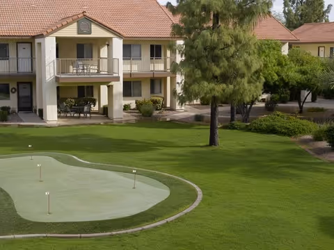 View of a senior living facility with a two-story building featuring balconies and patios. In the foreground, there is a small putting green surrounded by well-maintained grass and trees.