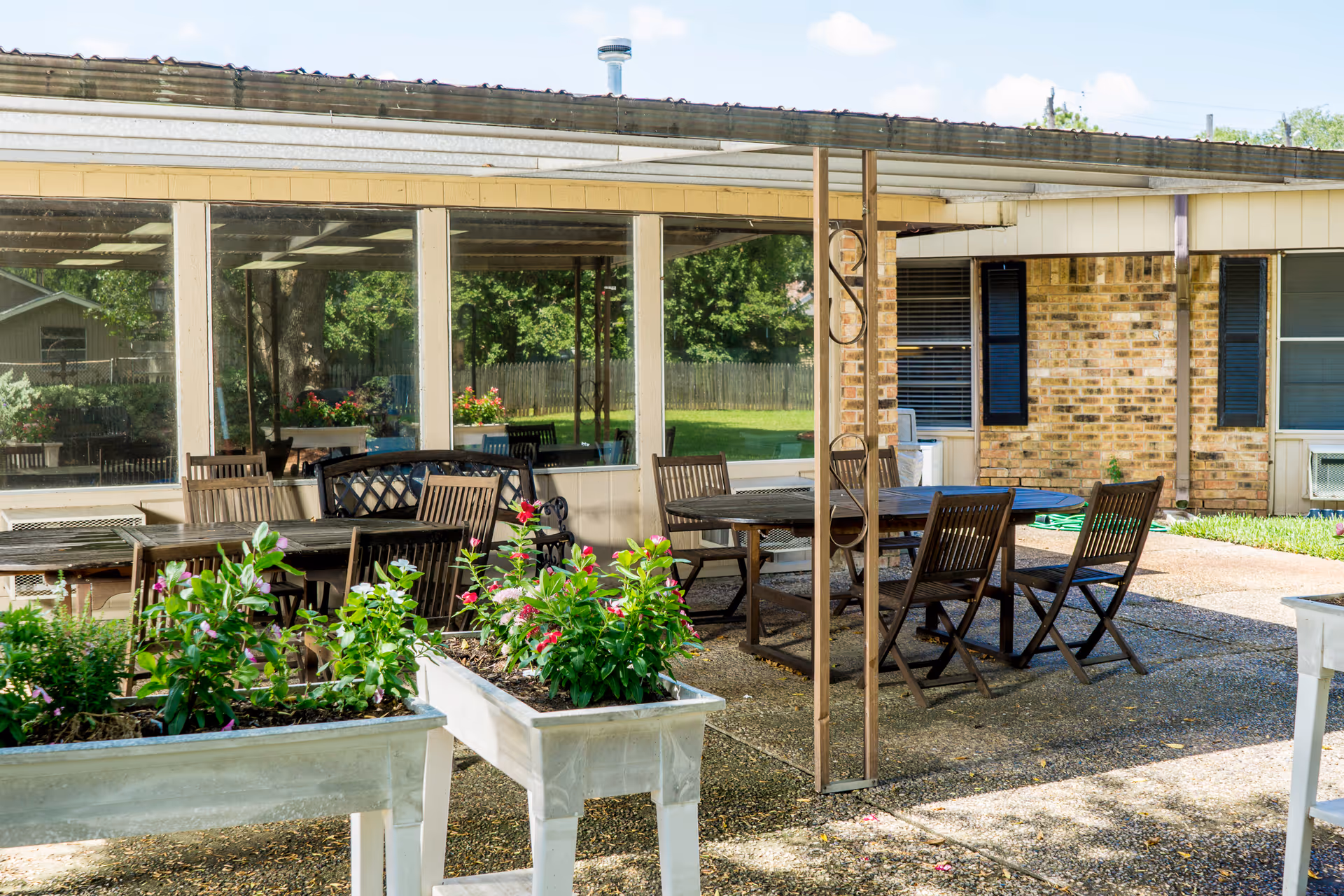 Outdoor patio area at Sugar Land Health Care Center with multiple wooden tables and chairs under a covered roof. Raised garden beds with green plants and flowers are in the foreground. The building exterior features brick walls and windows with blue shutters.