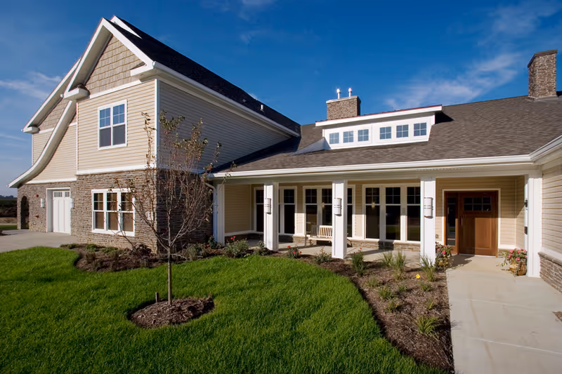 Exterior view of a large, modern residential building with beige siding, stone accents, and a covered porch with white columns. The building is surrounded by a well-maintained lawn and landscaping under a clear blue sky.