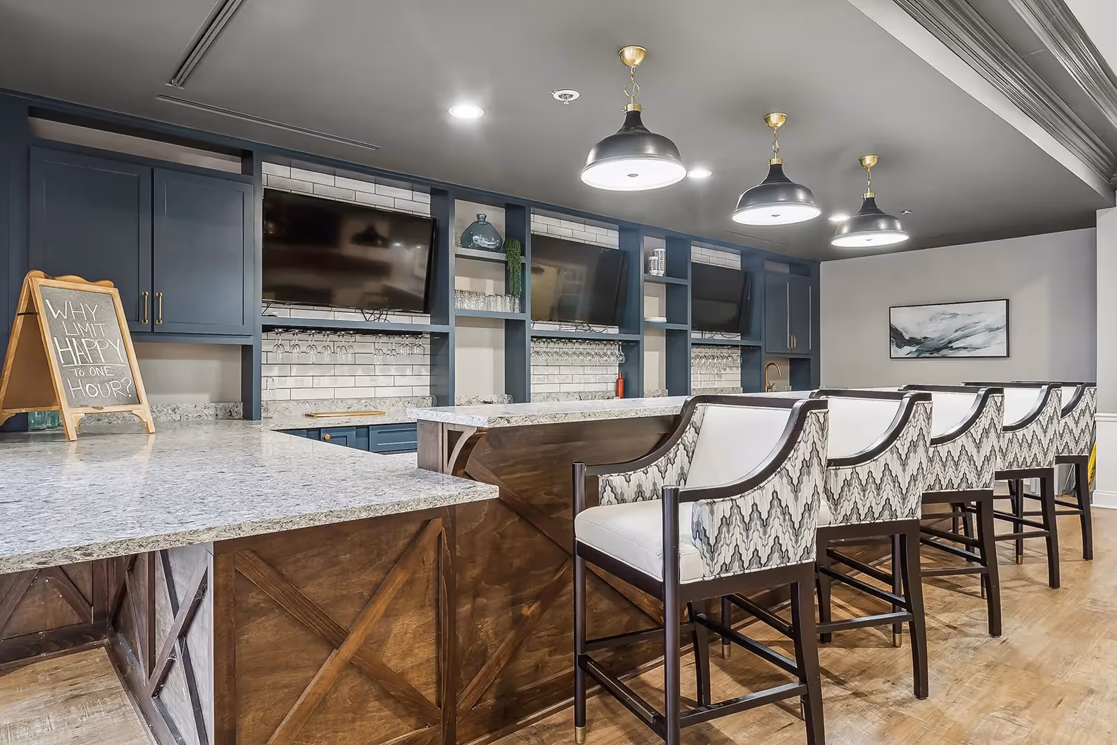 Communal bar-style seating area with a long granite countertop, patterned upholstered bar stools, pendant lights, and built-in shelving and TVs.
