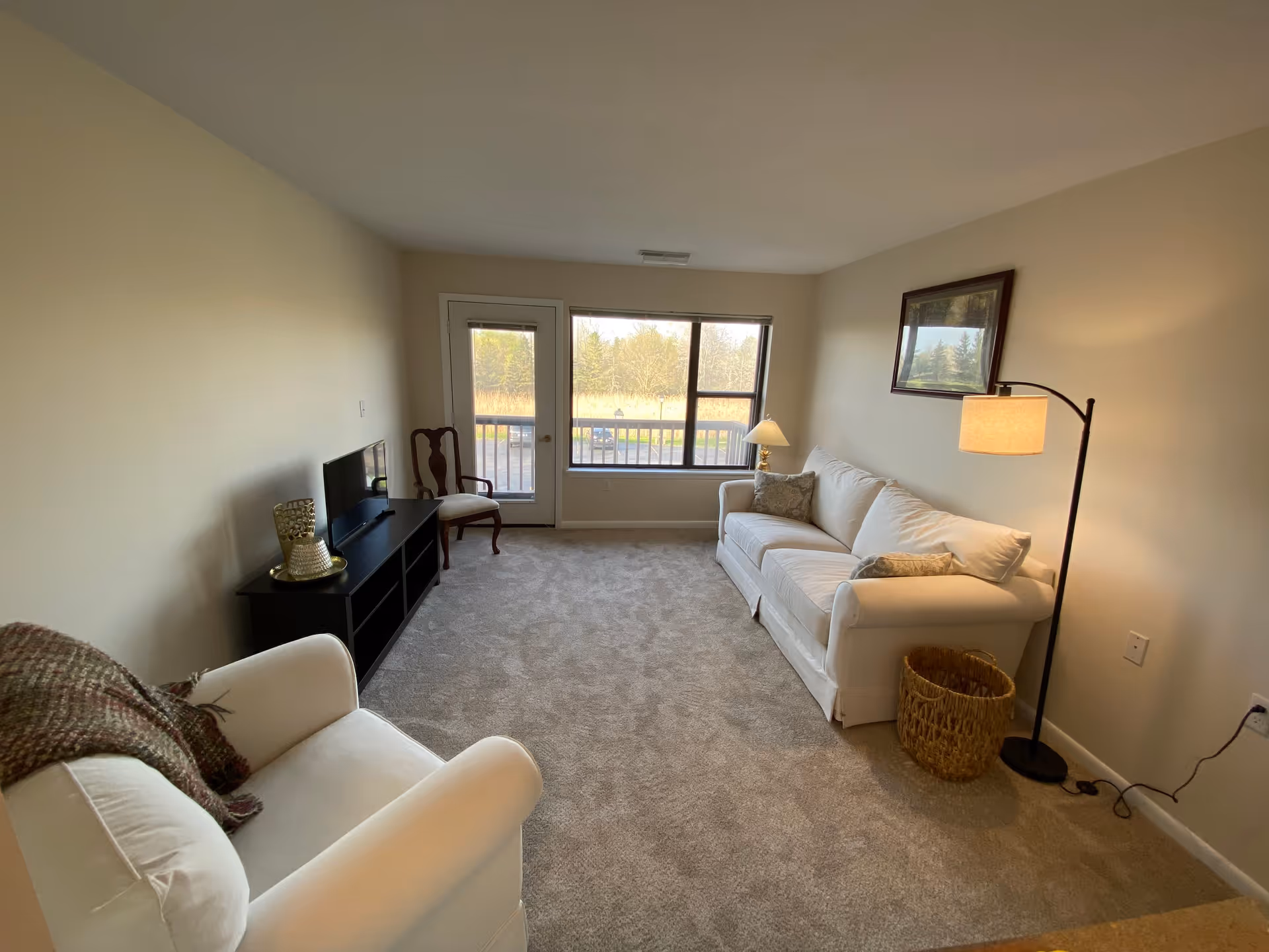 A cozy living room with beige walls and carpet. The room features a white sofa with two patterned pillows, a white armchair with a brown throw blanket, a black TV stand with a flat-screen TV, a wooden chair, a floor lamp, a table lamp on a small side table, a wicker basket, and a framed picture on the wall. A large window and a glass door provide a view of an outdoor area with trees.