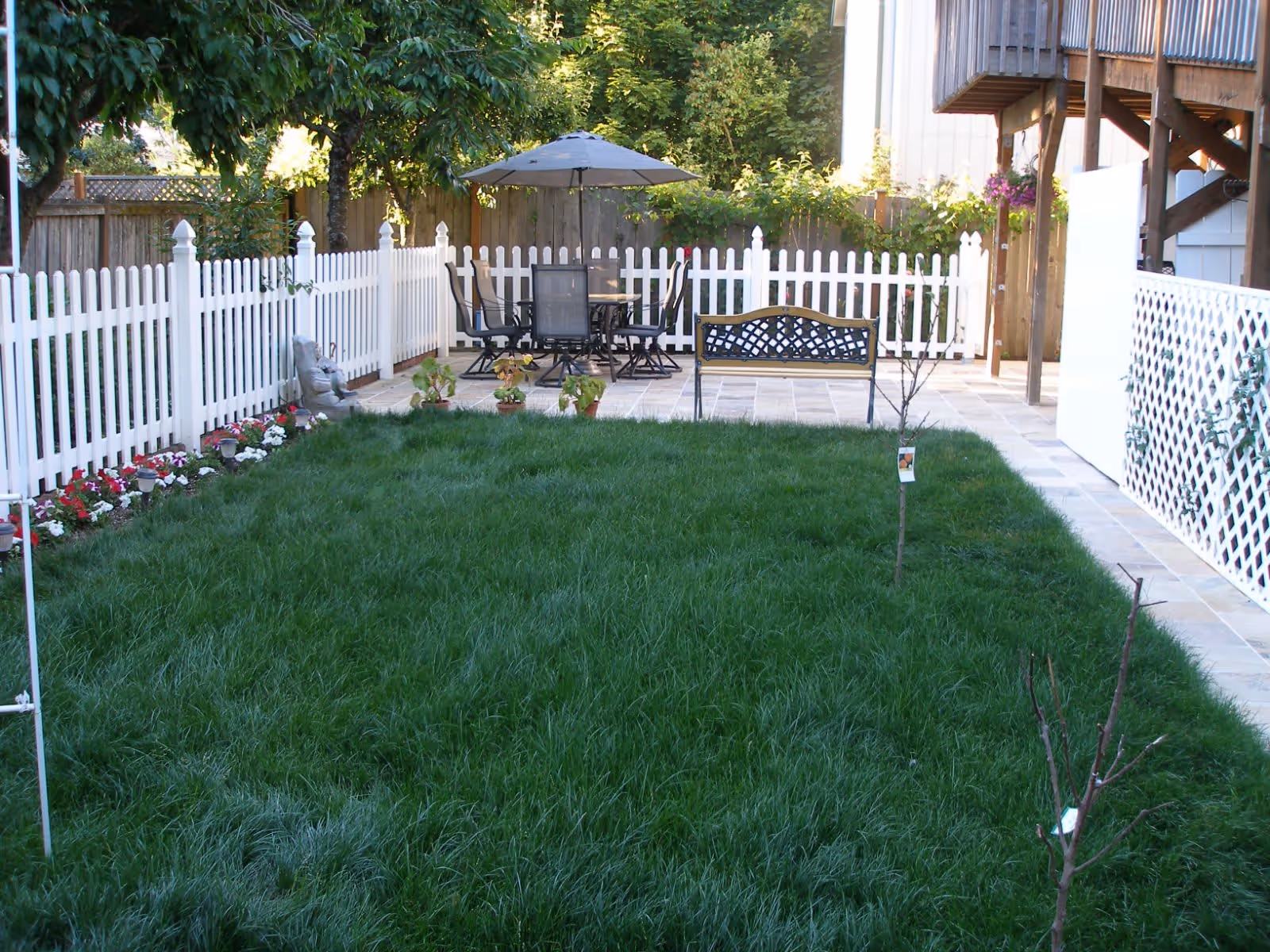 A fenced backyard garden area with green grass, a white picket fence, a small patio with a table, chairs, and an umbrella, a bench, and some small plants and flowers along the fence.