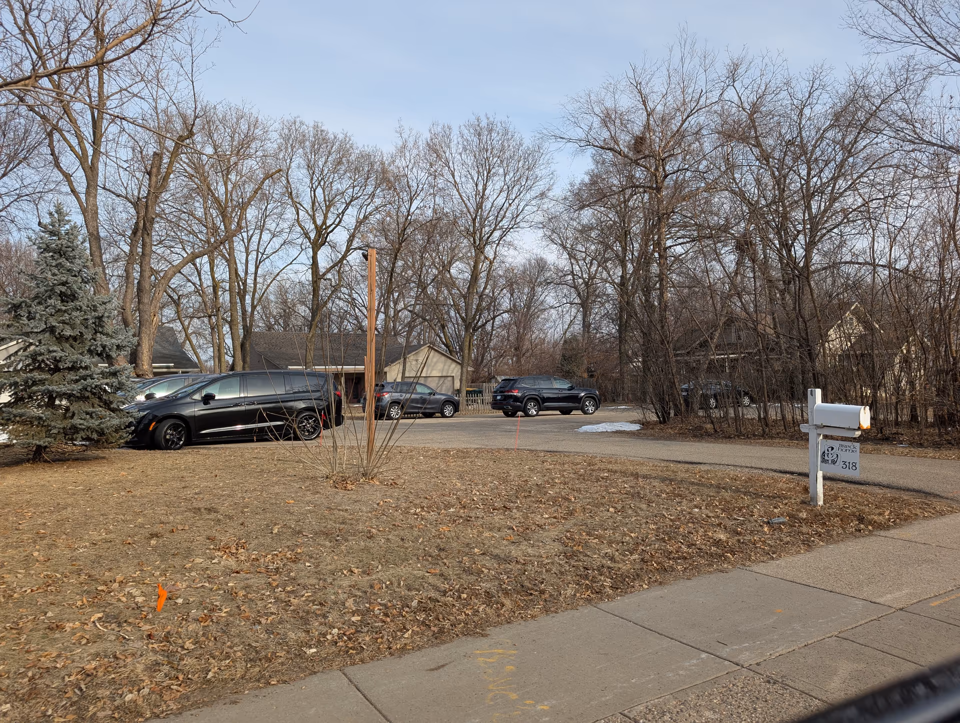 A parking area with several parked cars surrounded by leafless trees and a few houses in the background. There is a mailbox on the right side near the sidewalk with a sign that reads 'Breck Home Residential Care Center 318'. The ground is covered with dry grass and fallen leaves.