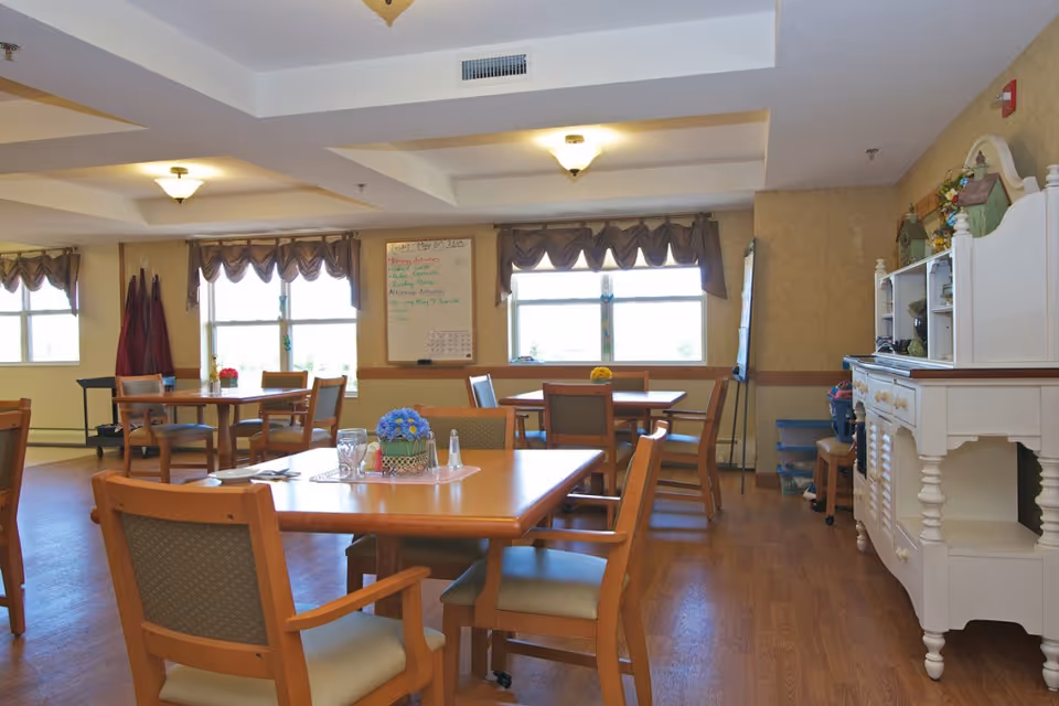 A bright dining room in a senior living facility with wooden tables and chairs arranged neatly. The room has large windows with valance curtains allowing natural light to fill the space. A whiteboard with notes is mounted on the wall, and a white sideboard cabinet with decorative items is placed against one wall. The floor is wooden, and the ceiling has recessed lighting fixtures.