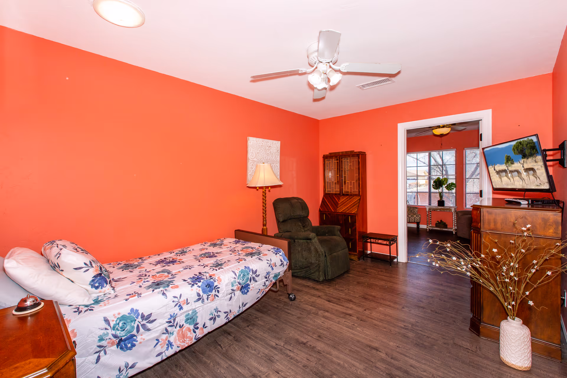 A coral-colored bedroom with a single bed with floral bedding, a recliner, dresser with TV, ceiling fan, and an open doorway to a sunlit sitting area.