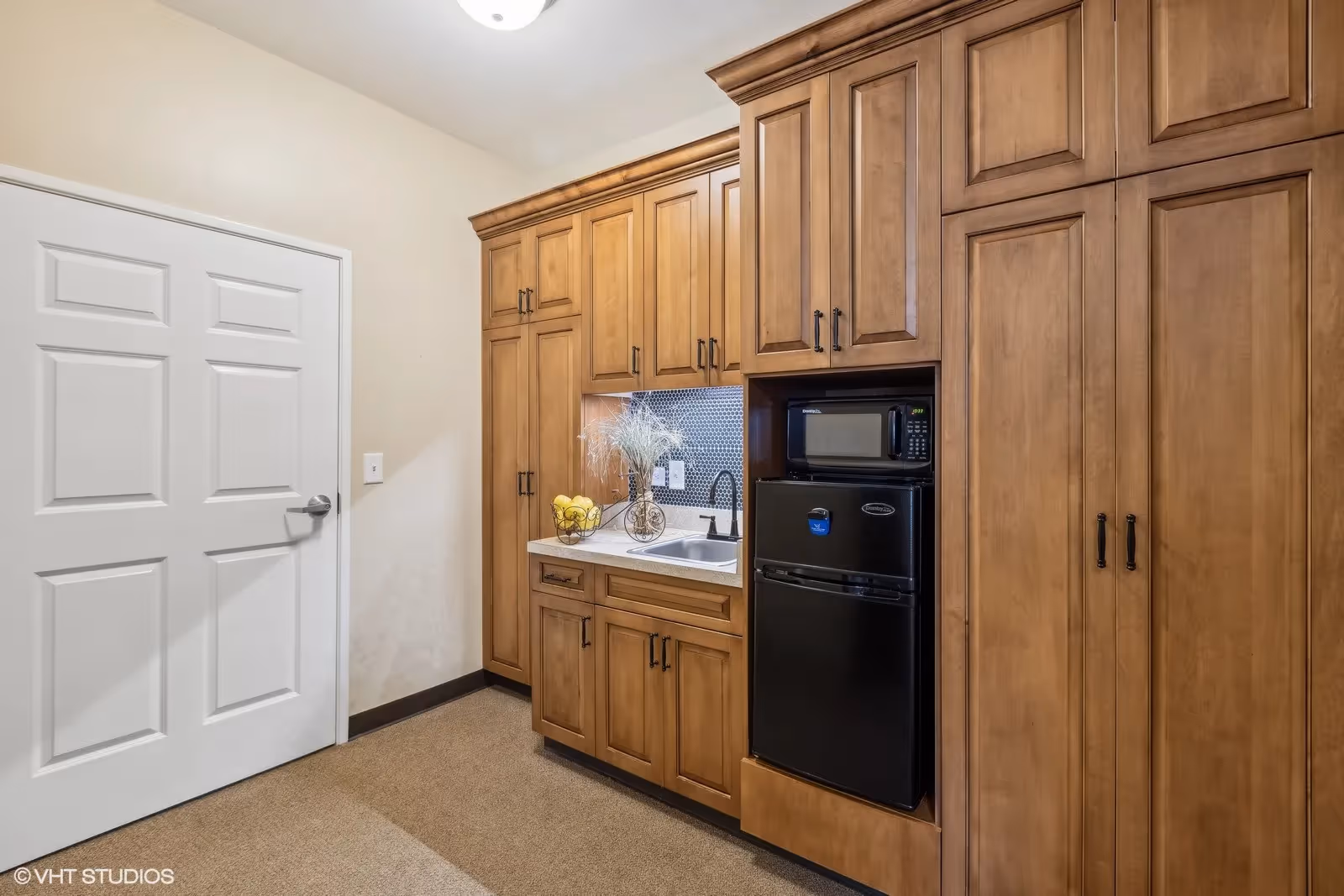 A compact kitchenette area featuring wooden cabinets, a small black refrigerator with a microwave on top, a white countertop with a sink, and decorative items including a bowl of lemons and a vase with dried flowers. There is a white door to the left and beige walls and carpeted floor.