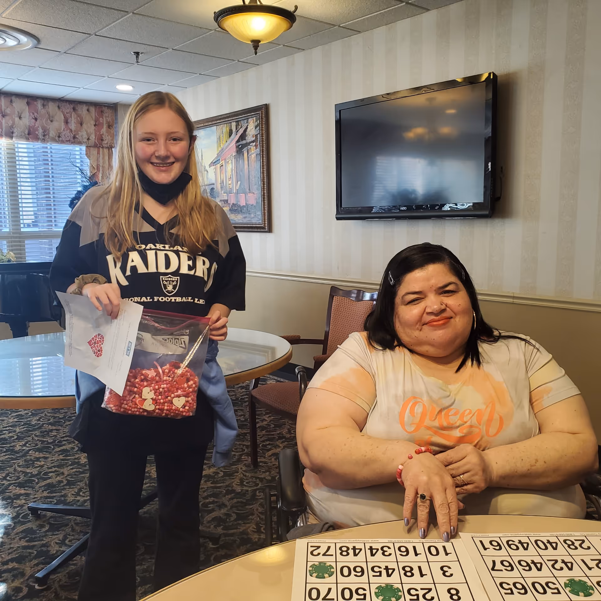 Two people in a senior living common room, one standing holding a bag of candy and papers and the other seated at a table with bingo cards.