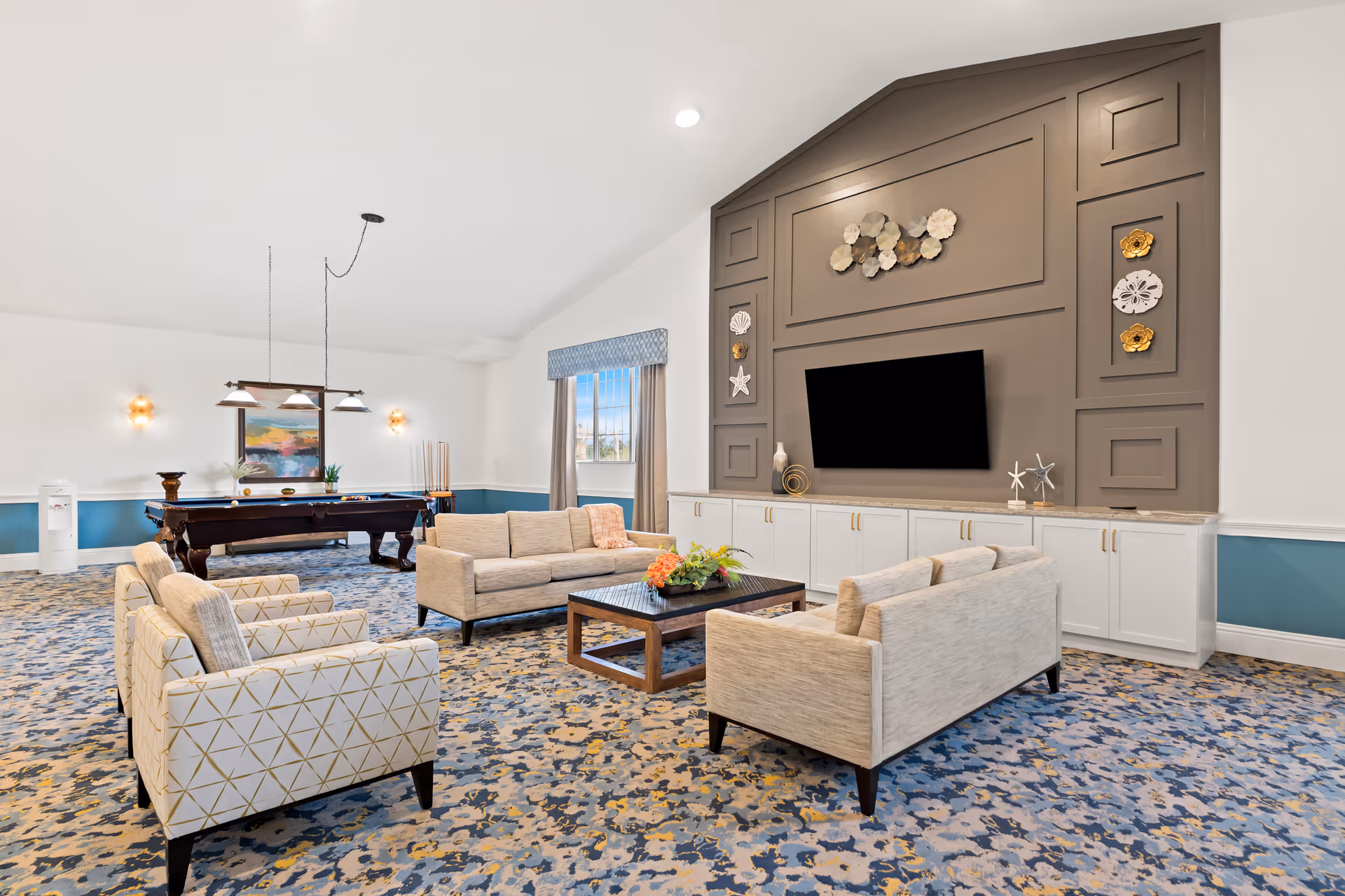 A spacious and well-lit living room area with two beige sofas and two patterned armchairs arranged around a dark wooden coffee table with a flower arrangement. The room features a large wall with decorative paneling, a mounted flat-screen TV, and coastal-themed wall decorations. In the background, there is a pool table with hanging lights above it, a window with curtains, and a water cooler.