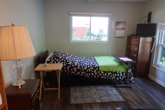 A small bedroom with a single bed covered with a black and white floral blanket and a green throw. There is a wooden nightstand with a glass lamp on the left side of the bed and a small wooden folding table next to the bed. A window above the bed shows an outside view of a brick building and greenery. On the right side of the room, there is a wooden dresser with a small TV on top and a calendar hanging on the wall. The floor is wooden with a small patterned rug in front of the bed.
