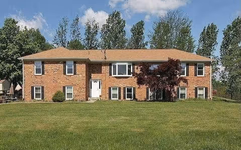 Front exterior view of a two-story brick building with a tan roof, multiple windows, a white front door, and a small tree in front. The building is surrounded by a large grassy lawn and trees in the background under a partly cloudy sky.