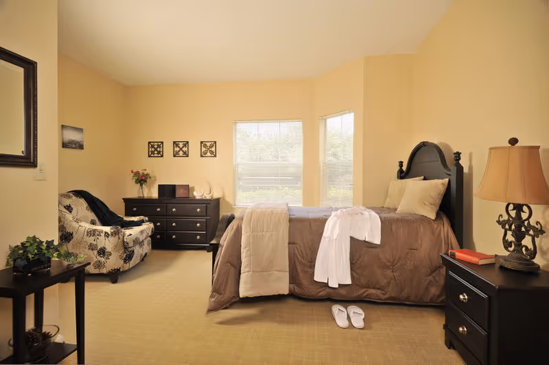 A cozy bedroom with a single bed covered in a brown quilt and beige blanket, a white robe draped over the bed, and slippers on the floor. The room features a dark wooden headboard, a nightstand with a decorative lamp and a book, a dresser with decorative items and flowers, a patterned armchair, and two windows letting in natural light.