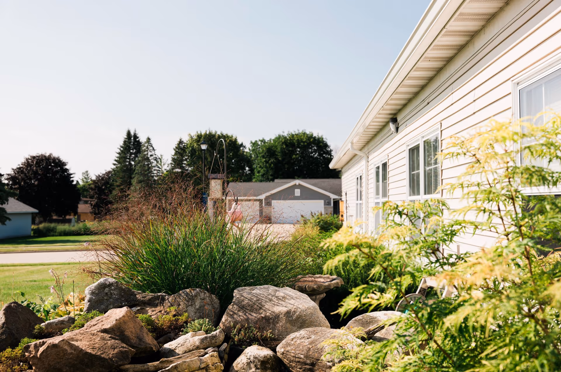 Landscaped yard with rocks, grasses, and shrubs beside a light-colored single-story building under a clear sky.