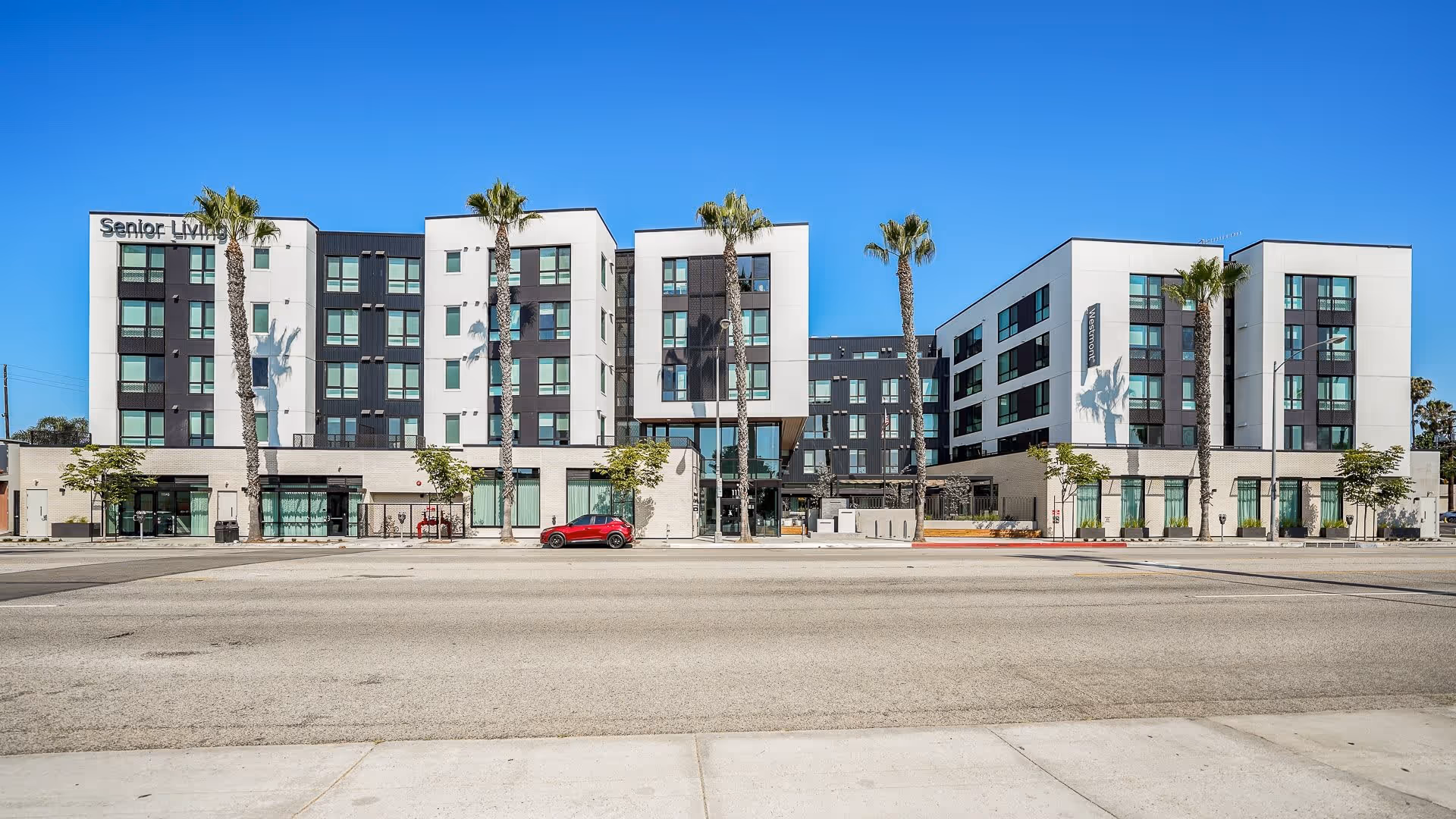 Front view of a modern multi-story senior living building with palm trees along the sidewalk and a red car parked on the street under a clear blue sky.